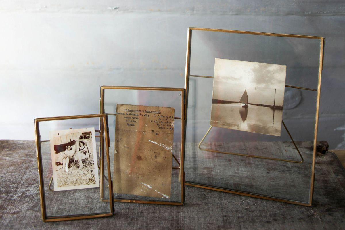 Three elegant Danta Antique Brass Frames by nkuku of varying sizes stand on a textured surface. The left displays a sepia photo of a woman, the middle holds a faded note, and the right shows a vintage sailboat on calm water.
