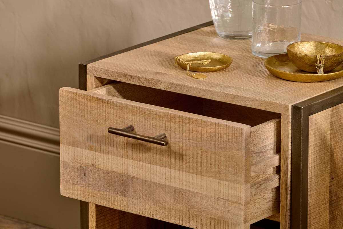 The Dasai Mango Wood Bedside Table by nkuku, featuring an open drawer with a simple metal handle, displays two gold bowls with jewelry and two clear glasses on top. Soft lighting highlights its handcrafted mango wood grain against a beige wall.
