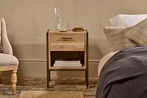 The Dasai Mango Wood Bedside Table by nkuku, crafted from mango wood with an iron frame, includes a drawer and open shelf for books. A glass carafe, drinking glass, and two golden dishes sit atop it beside textured bedding and a cushioned chair.