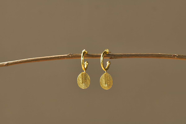 The Esta Hoop Earrings - Gold by nkuku feature hammered oval gold charms, hanging from a thin branch against a warm beige background. Their textured surface shines in the light, showcasing an elegant and minimal design. One size fits all.