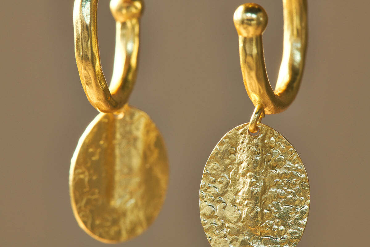 Close-up of nkuku’s Esta Hoop Earrings in gold, featuring textured, hammered oval pendants. The softly blurred background highlights the artisanal appeal and unique beauty of this handcrafted jewelry. One size.