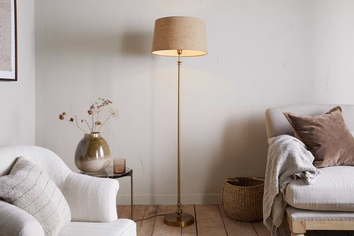 A cozy living room with a beige armchair and sofa, a nkuku Ehadi Antique Floor Lamp in antique brass, a woven basket, side table with ceramic vase of dried flowers, and a soft throw. Warm neutrals create a calm, inviting atmosphere.
