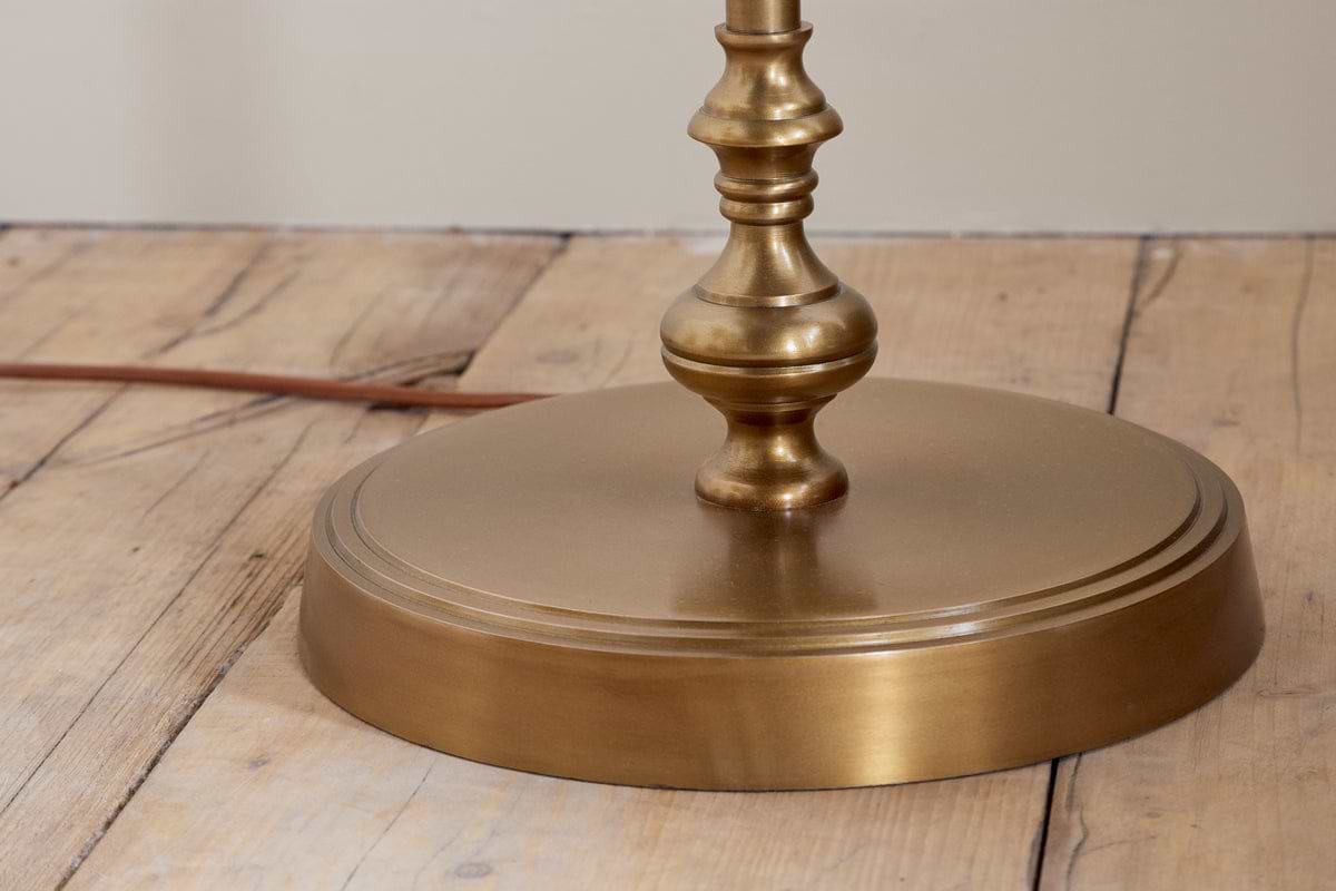 The nkuku Ehadi Antique Floor Lamp in antique brass features a rounded, stepped base with a warm golden finish, set on a light wooden floor. An electrical cord extends from behind the lamp.