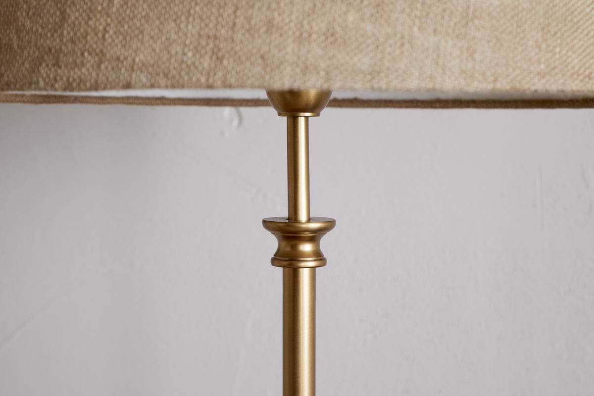 A close-up of the nkuku Ehadi Antique Floor Lamp in antique brass, featuring a slender metallic top and decorative collar with a textured beige shade, set against a plain white wall for a minimalist look.