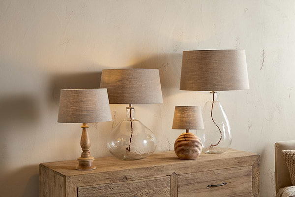 Four Embaata Linen Lampshade table lamps by nkuku, featuring natural textured linen shades and bases in wood, glass, and ceramic, are displayed on a rustic sideboard against a beige wall—ideal as bedroom lighting.