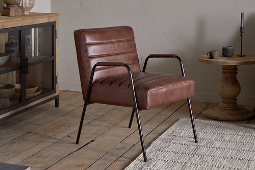The Hadithi Leather Occasional Chair in chocolate brown by nkuku, featuring black metal legs, is placed on a wooden floor beside a woven rug. A glass-door wooden cabinet and round side table with candle and cup enhance the cozy space.