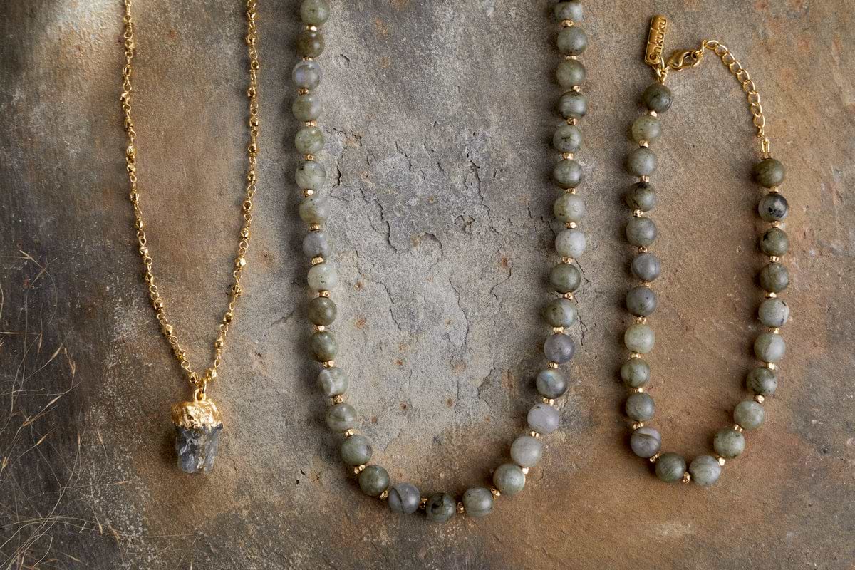 Three pieces of jewelry on a textured surface: a gold chain pendant necklace with a crystal, the nkuku Hadari Labradorite Necklace in gold with round grey-green stones, and a matching beaded bracelet with a gold clasp.