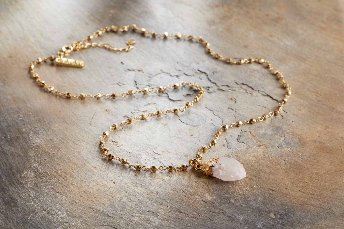 The nkuku Hadari Rose Quartz Necklace features a delicate gold chain with subtle beads and a rough teardrop-shaped rose quartz pendant, elegantly displayed on textured light brown stone—perfect for any occasion.