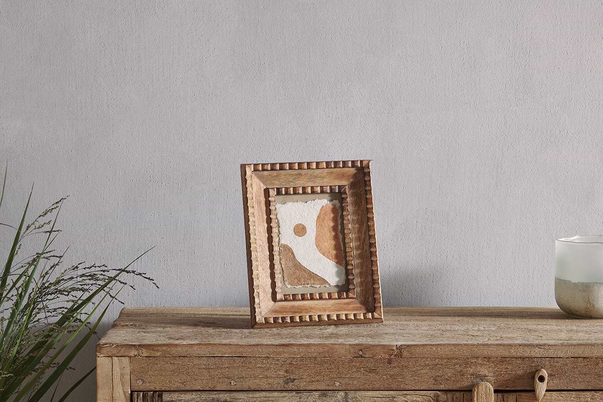 A wooden cabinet displays the nkuku Hansika Mango Wood Frame - Natural, showcasing abstract brown, white, and tan art. A green plant sits on the left and a white candle in a glass holder on the right, with a light gray wall behind.