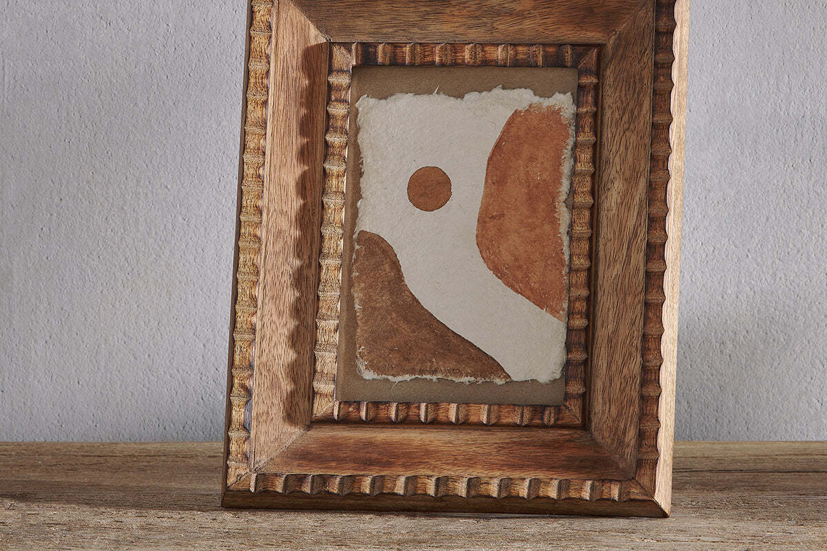 The nkuku Hansika Mango Wood Frame – Natural holds abstract art with curved brown shapes and a circle on textured paper, set against a white background. The rustic, hand-carved textured frame rests on wood with a plain gray wall behind.