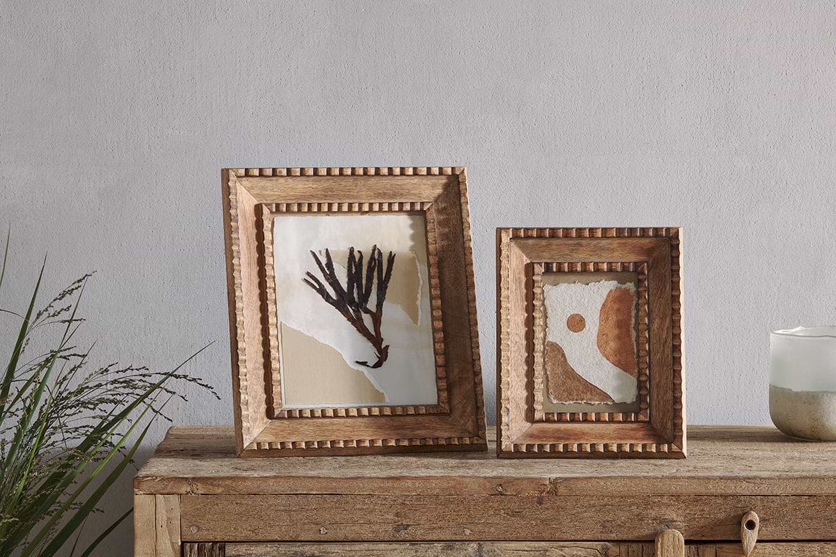 Two nkuku Hansika Mango Wood Frames in natural finish, one with beaded edges, display abstract and earth-toned art on a rustic table. Nearby sit a glass candle and green plant, all set against a pale gray wall.