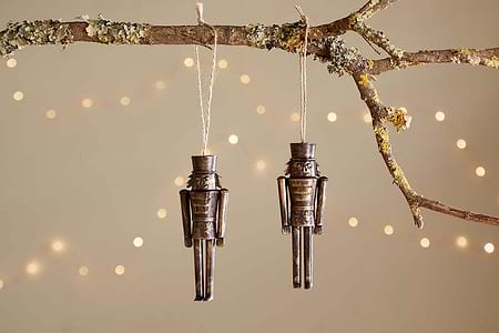Two Imay Nutcracker Baubles in antique brass by nkuku hang from twine on a mossy branch, with warm white string lights softly glowing in the background—perfect for adding vintage charm to your Christmas tree décor.