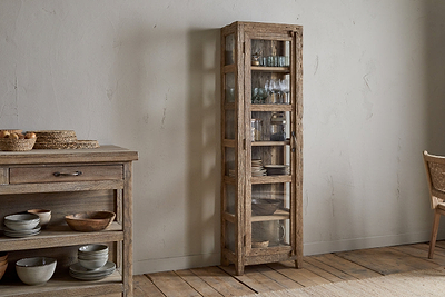 The nkuku Ibo Reclaimed Wood & Glass Cabinet - Natural - Tall, with antique brass handles, showcases glassware and bowls in a cozy room featuring a wooden floor, sideboard with dishes, and a wicker chair.