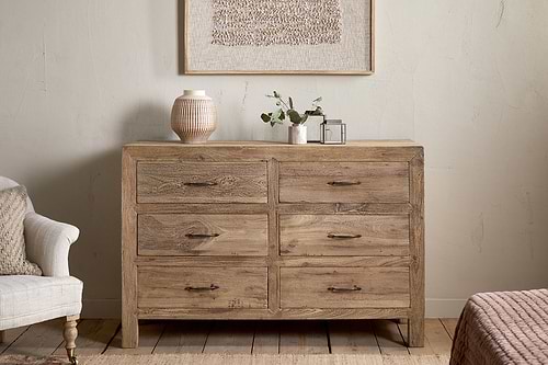 The nkuku Iya Reclaimed Wood Chest of Drawers - Natural - Large, featuring six drawers, stands against a light wall, topped with a textured vase, small plant, and clear box. An armchair and bed are partially visible on either side.