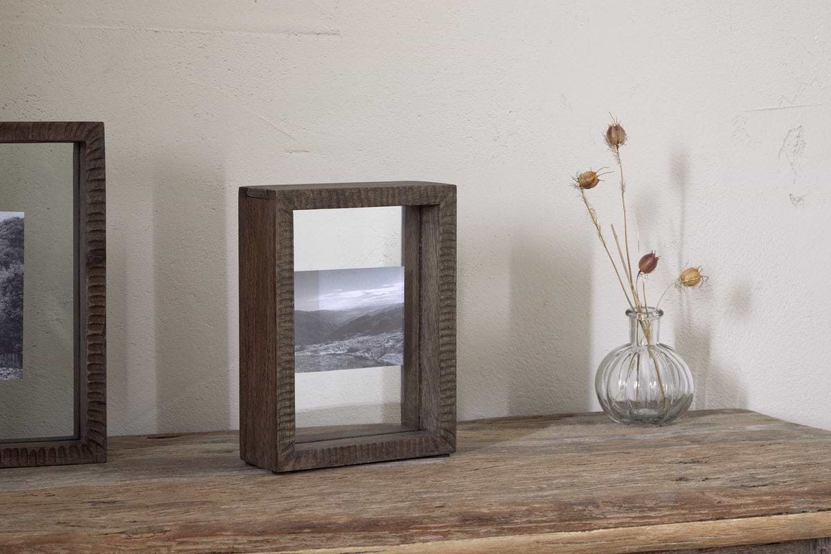 A black-and-white landscape photo is displayed in the nkuku Indu Standing Mango Wood Frame - Dark Ash, set on a wooden table beside a small glass vase with dried stems, all against a plain light-colored wall.