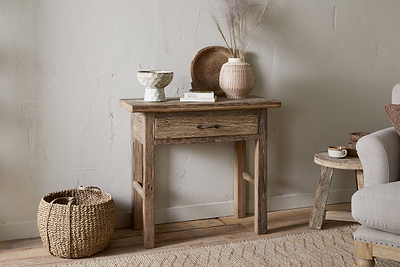 The nkuku Ibo Reclaimed Wood 1 Drawer Console Table in Natural brings rustic charm to your entryway, styled with a ceramic bowl, book, wooden tray, and vase with dried grasses. A woven basket sits nearby next to a cozy gray armchair.