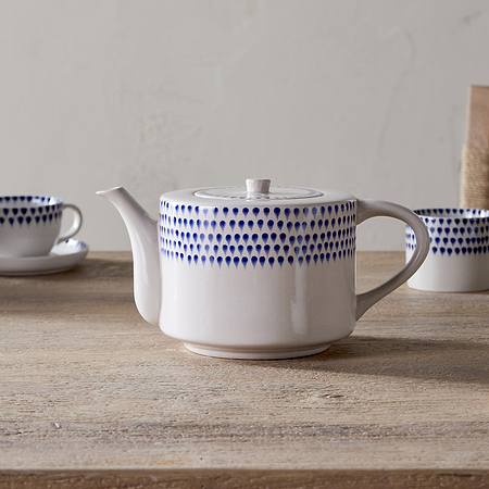 The nkuku Indigo Drop 4 Person Teapot in Cream & Indigo, adorned with hand-painted blue dots, sits on a wooden table with matching teacups and saucers—an ideal gift for tea lovers. A woven chair appears in the background.