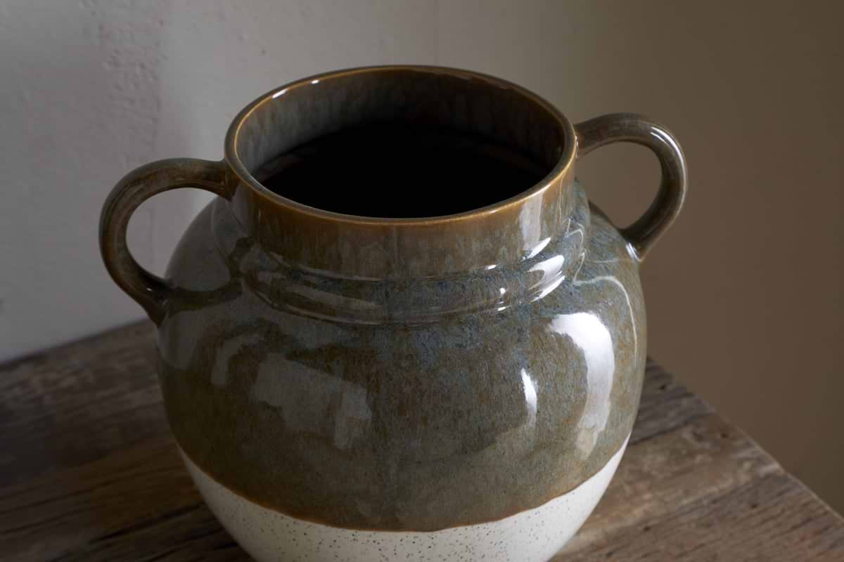 The nkuku Inha Reactive Glaze Vase in Dark Olive & Natural features two handles and sits on a rustic wooden surface, showcasing a glossy dark olive upper half, a matte speckled white base, and smooth texture highlighted by light and shadow.