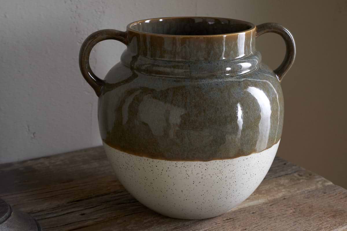 The Inha Reactive Glaze Vase by nkuku sits on a wooden surface, featuring two handles. Its top half is glossy dark olive, while the speckled matte bottom is natural off-white, with reflections and shadows enhancing its smooth texture.
