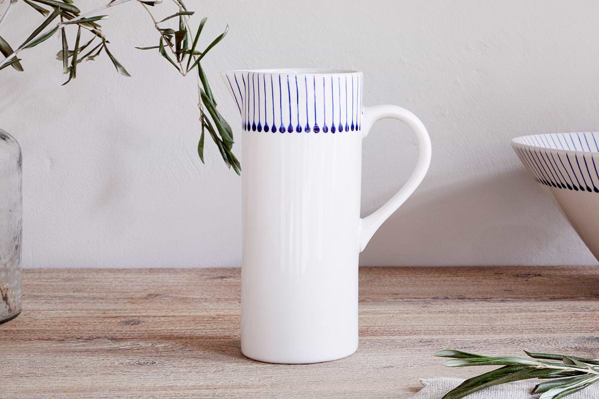 The Iba Jug - Indigo by nkuku, a tall white ceramic jug with vertical blue lines and dotted details near the rim, rests on a wooden surface beside olive branches and a matching bowl in a minimal, natural setting.