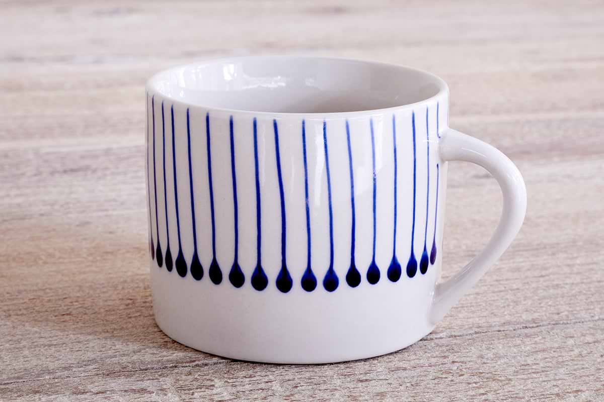 The nkuku Iba Mug - Indigo - Small (Set of 2) features a white ceramic design with a simple handle and hand-painted vertical blue teardrop lines—ideal for fans of unique, artisanal coffee cups.