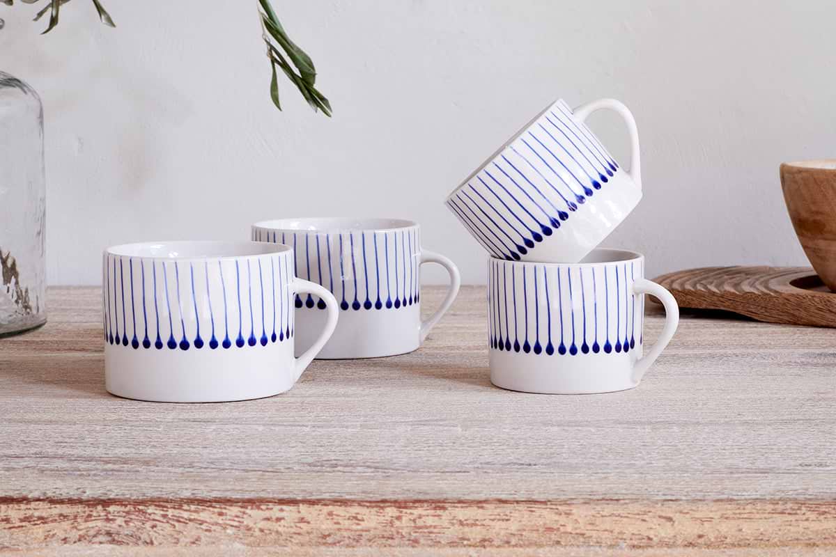 Two sets of nkuku Iba Mug - Indigo - Large (Set of 2) feature white mugs with vertical blue lines and teardrop designs, arranged on a light wooden table; three stand upright, one rests at an angle, with glass bottle and bowl in the background.