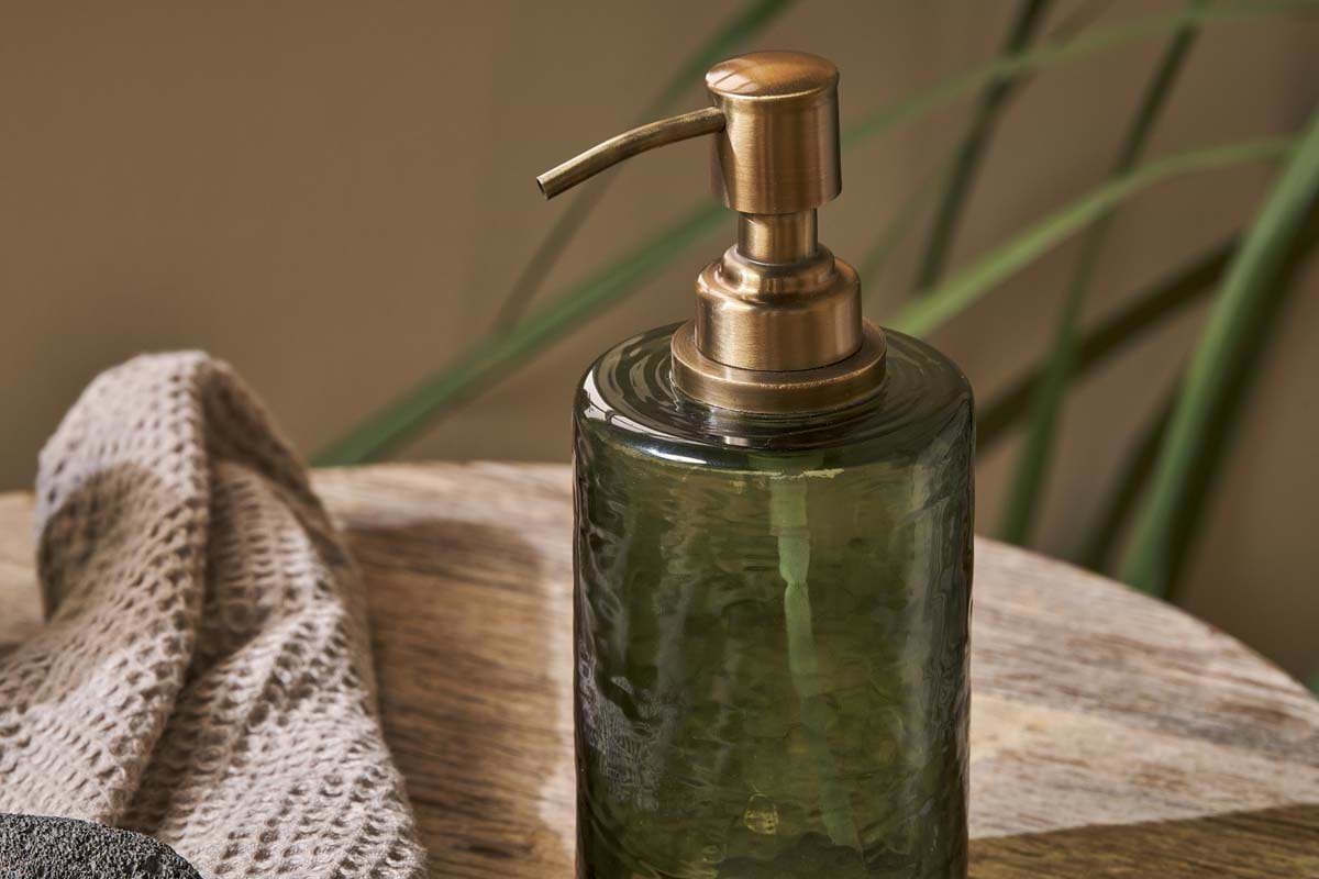 The Ilcoso Recycled Hammered Glass Soap Dispenser in green by nkuku rests on wood, with a textured beige towel nearby and soft green leaves in the background, creating a serene spa-like scene.