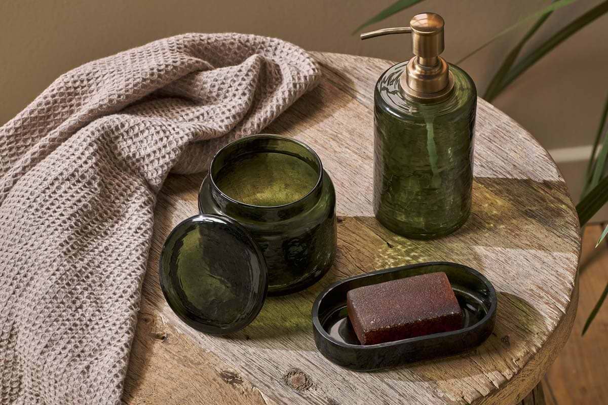 The nkuku Ilcoso Recycled Hammered Glass Soap Dispenser in green sits on a rustic wooden table, accompanied by a beige towel, a lidded green glass jar, and a soap dish with brown soap. Green leaves partly frame the image's right edge.
