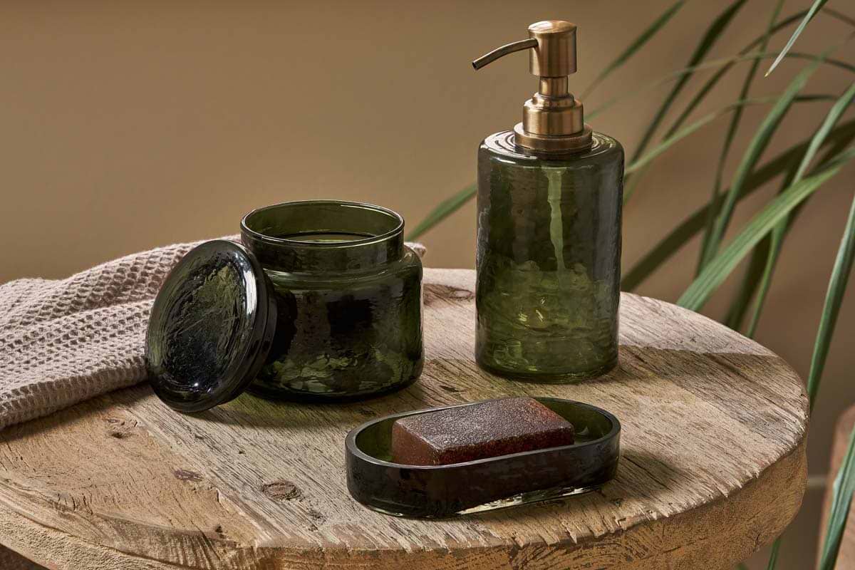 A rustic wooden table displays nkuku’s Ilcoso Recycled Hammered Glass Soap Dispenser in green, paired with matching lidded jar and soap dish. A beige towel and tall greenery enhance the natural, spa-inspired scene.