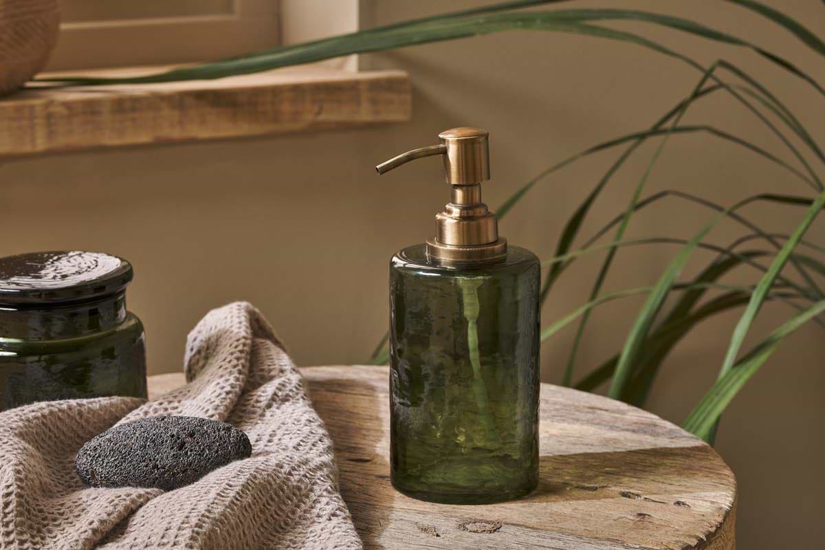 The Ilcoso Recycled Hammered Glass Soap Dispenser in green by nkuku, featuring a brass pump, sits on a rustic wooden table beside a gray towel and pumice stone, with greenery and a glass jar softly lit in the background.