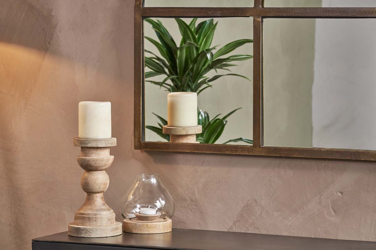A beige wall features a black table topped with two wooden candle holders, cream candles, and a glass tealight holder. Above hangs the nkuku Imoma Iron Overmantel Arch Mirror in brass, reflecting green foliage in warm, cozy light.