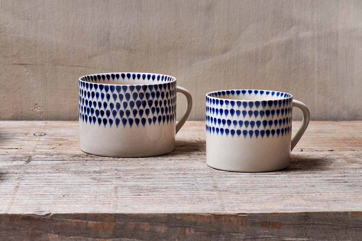 The nkuku Indigo Drop Mug - Cream & Indigo - Large (Set of 2) features handmade ceramic mugs with indigo teardrop patterns near the rim, displayed on a rustic wooden surface against a neutral textured background.