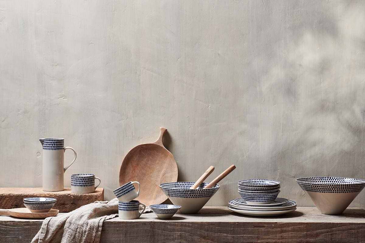 The nkuku Indigo Drop Mug - Cream & Indigo - Large (Set of 2) features white ceramic with blue dotted patterns, elegantly displayed on rustic wood with a beige cloth in the foreground and neutral background.