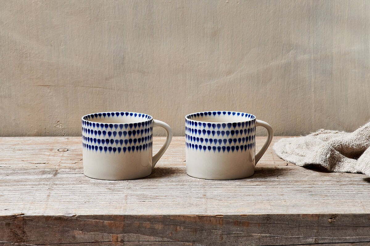 The nkuku Indigo Drop Mug - Cream & Indigo - Small (Set of 2) features hand-painted indigo designs on white ceramic, displayed side by side on rustic wood with a beige cloth for a minimalist, cozy look.