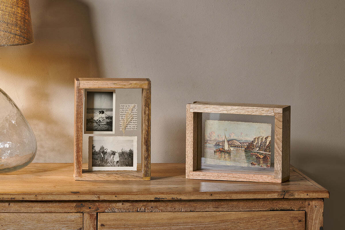 Two Indu Standing Mango Wood Frames by nkuku sit on a wooden surface. The left frame holds three black-and-white photos and a note; the right one displays a colorful riverside painting. A glass lamp is partially visible to the left.