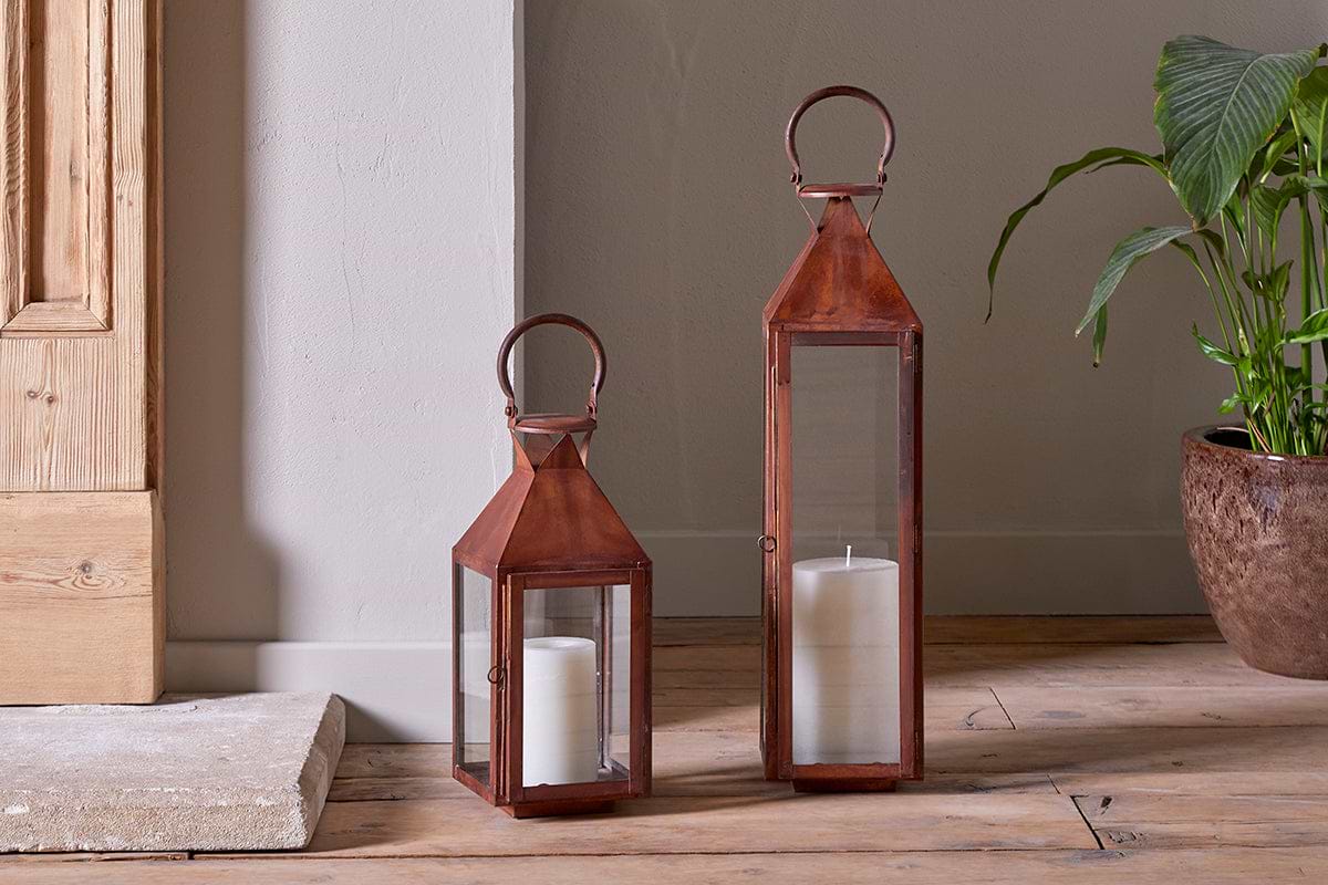 Two nkuku Isko Lanterns in a rich rust finish, each with glass panels and a white pillar candle, stand on wooden flooring near a beige wall. A green potted plant and wooden door add charm—perfect for cozy garden seating areas.