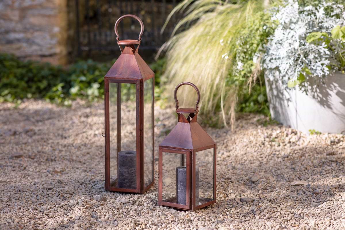 Two nkuku Isko Lanterns in rust, each with glass panels and handles, hold dark pillar candles on an outdoor gravel surface. Behind them are green plants, a large gray planter, garden seating, and a stone wall.