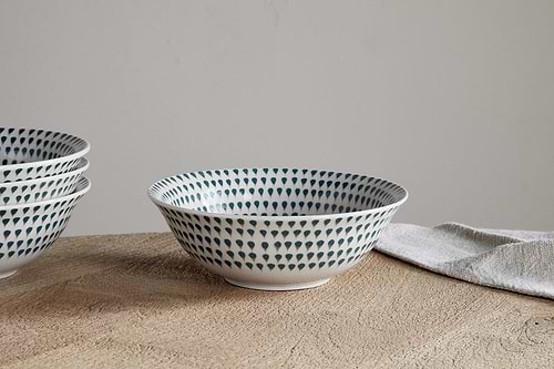 A white ceramic Juniper Drop Cereal Bowl in cream and juniper by nkuku, featuring a blue dotted pattern, sits on a wooden surface beside a folded beige cloth; more matching bowls are partially visible to the left—perfect for everyday dining.