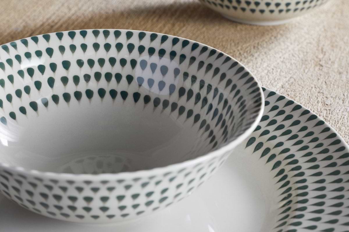 A close-up of the nkuku Juniper Drop Cereal Bowl in Cream & Juniper, featuring a green drop pattern on the rim, perfect for everyday dining, resting on a beige surface with another bowl partially visible in the background.