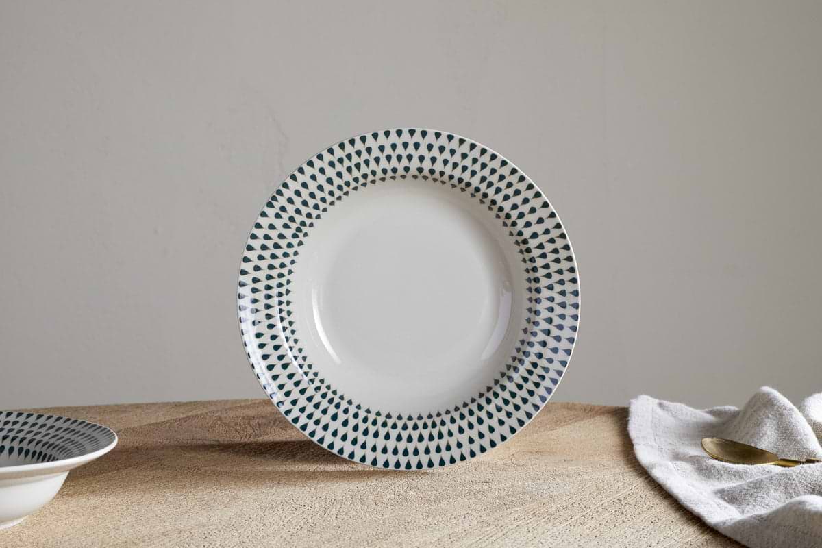 The Juniper Drop Pasta Bowl - Cream & Juniper by nkuku, a white ceramic bowl with small dark green drops around the rim, rests on a wooden surface with a cloth napkin and brass spoon partially visible to the right.