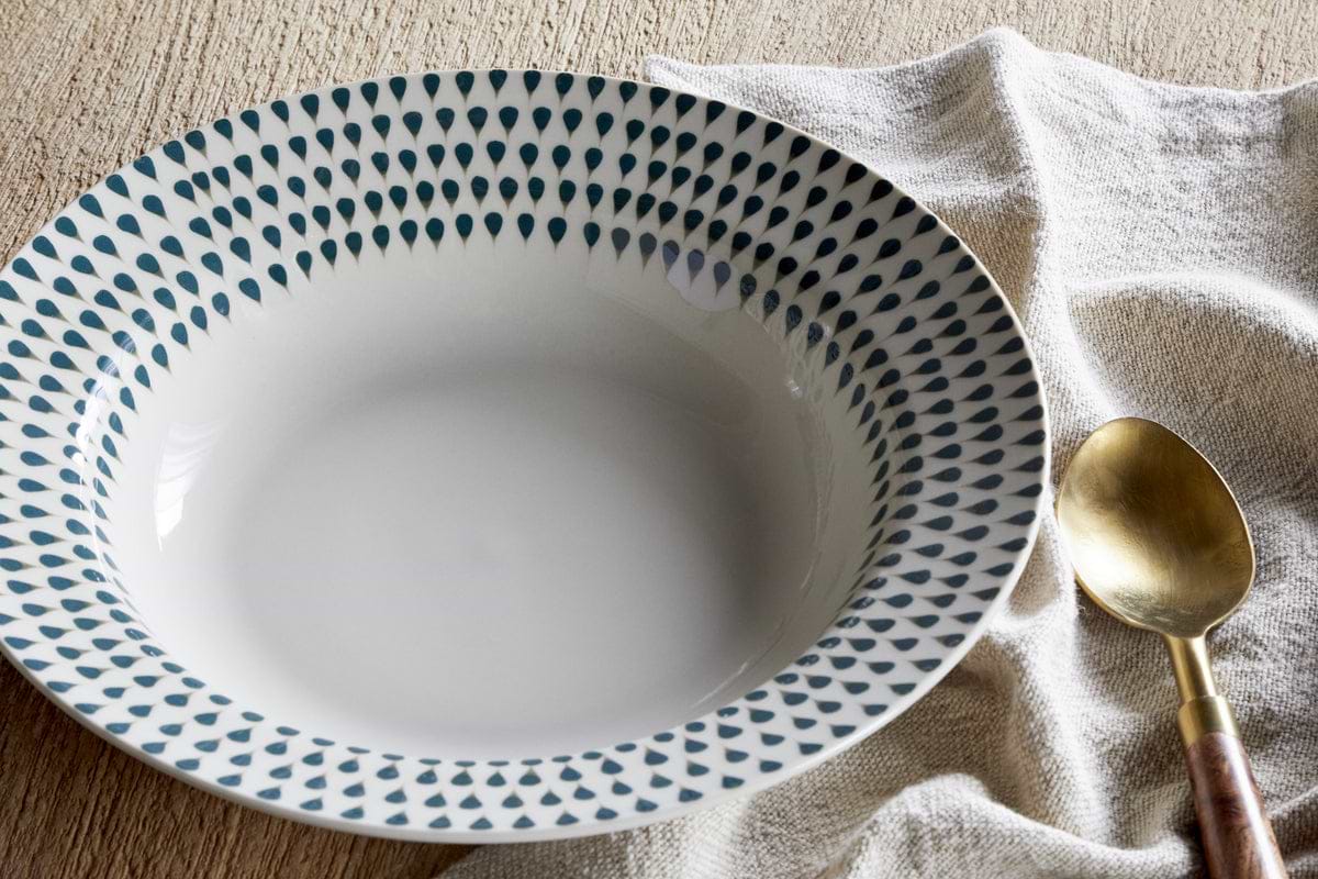 The Juniper Drop Pasta Bowl - Cream & Juniper by nkuku, featuring a blue teardrop rim design, sits on a beige cloth beside a gold spoon with wooden handle, highlighting contemporary tableware on a light wooden surface.