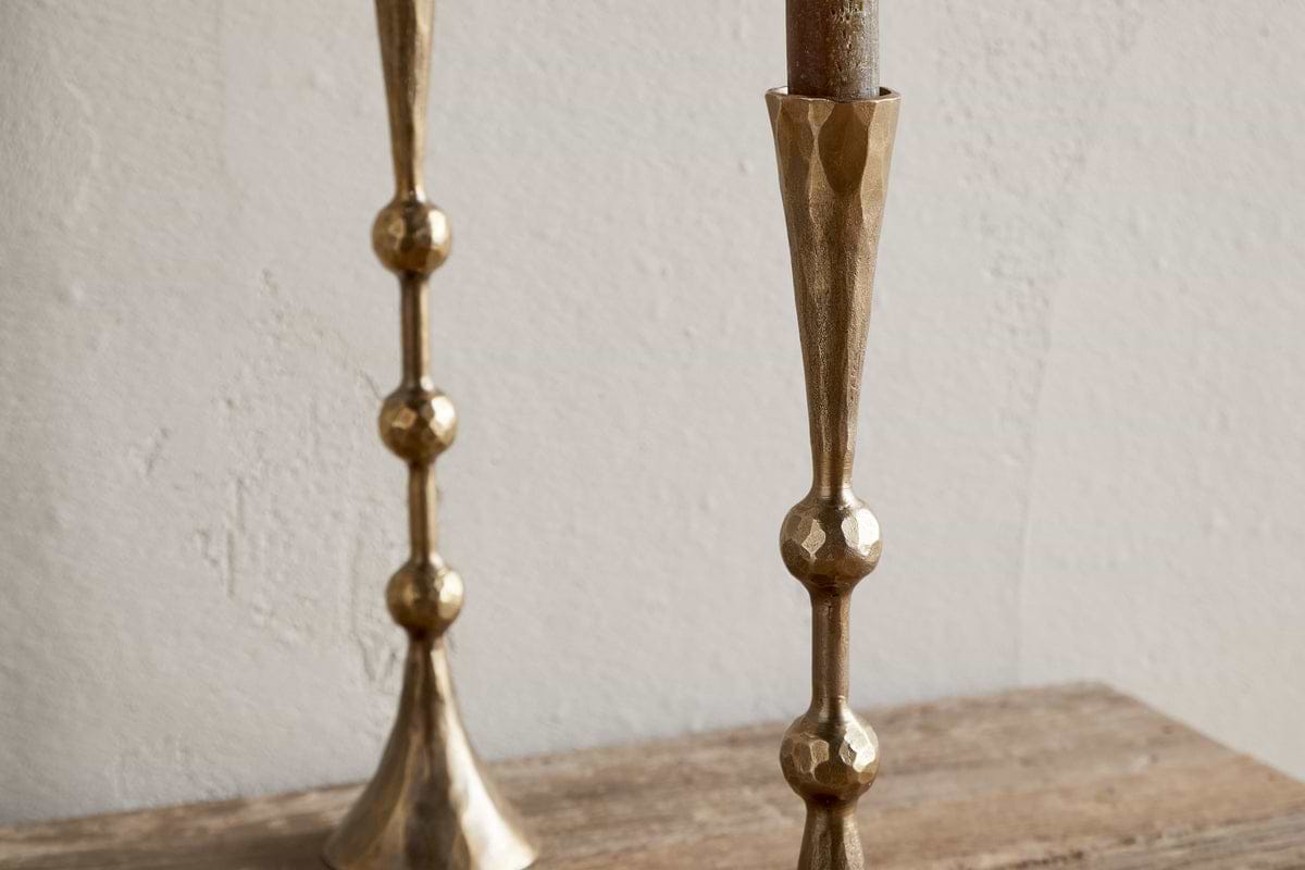 Two nkuku Javanee Candle Holders in antique brass, featuring a geometric hammered texture and spherical accents, stand on a rustic wooden surface with one partially visible on the left against a white textured wall.