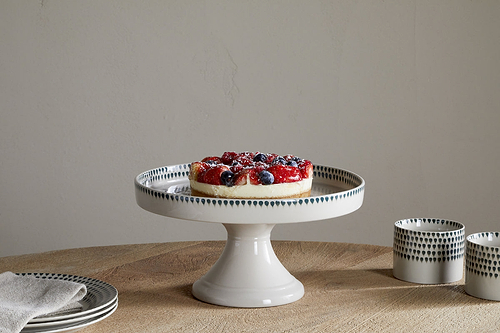A fruit tart topped with strawberries and blueberries rests on the nkuku Juniper Drop Ceramic Stand - Cream & Juniper, set on a wooden table with a stack of plates, a folded napkin, and two patterned cups nearby.