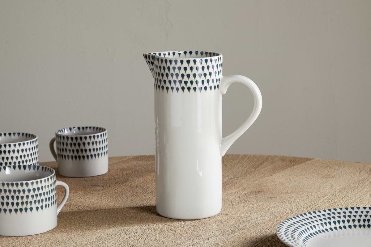 The nkuku Juniper Drop Jug - Cream & Juniper - Large is a white ceramic jug with a curved handle and blue teardrop pattern near the rim, shown on a wooden table next to matching cups and a plate.