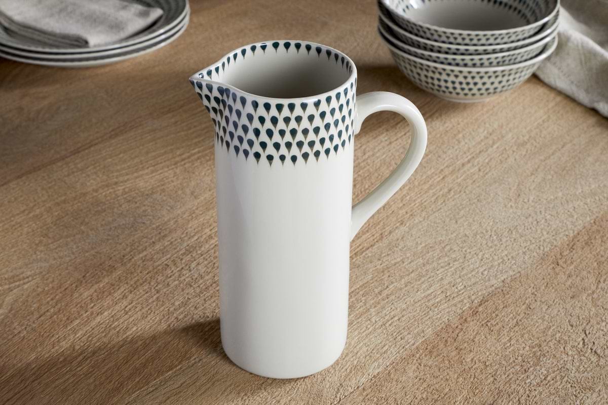 The nkuku Juniper Drop Jug – Cream & Juniper – Large, a white ceramic jug with a handle and blue teardrop accents near the rim, sits on a wooden table among stacked matching bowls and plates.