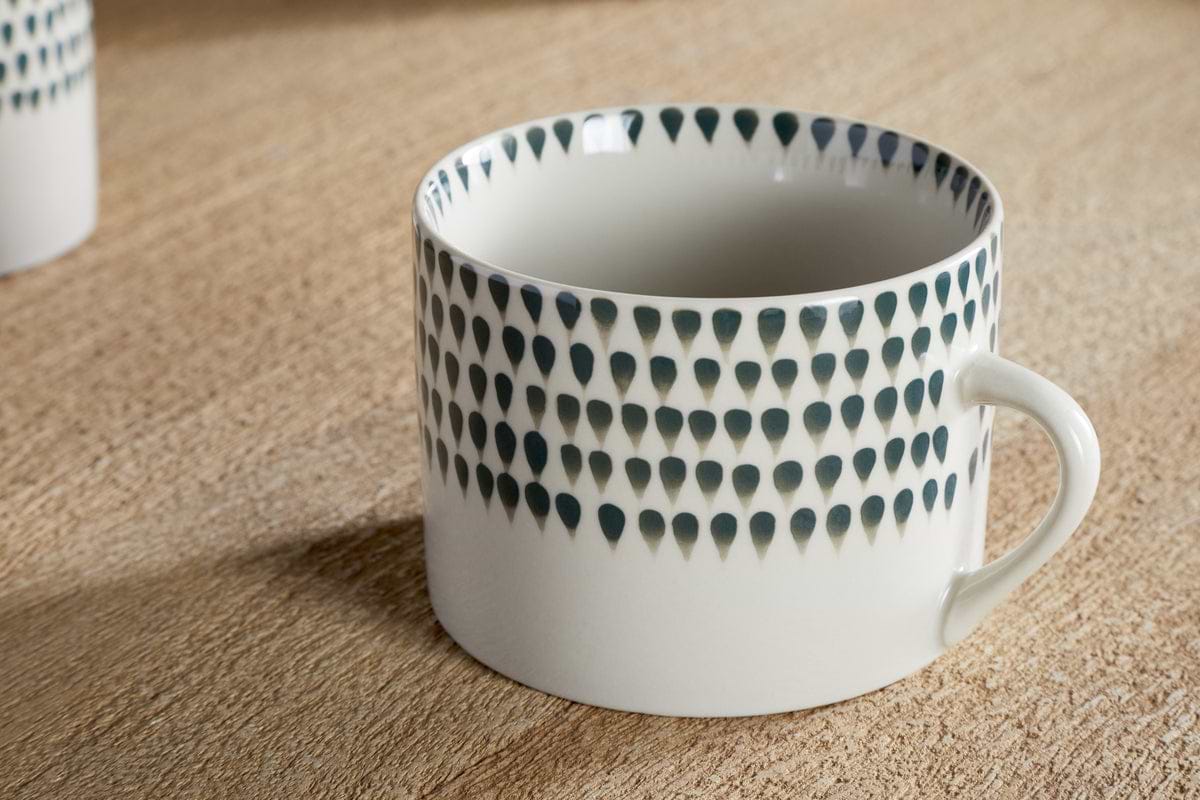 The nkuku Juniper Drop Mug - Cream & Juniper - Large (Set of 2) features a white ceramic body, dark blue teardrop pattern near the rim, simple handle, and glossy finish, shown on a wooden surface.