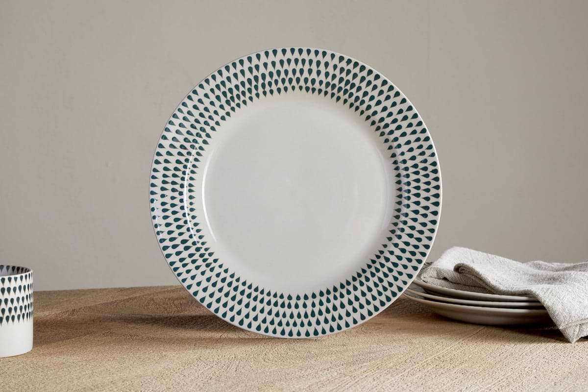 The nkuku Juniper Drop Dinnerware Set - Cream & Juniper (Set of 12) features white ceramic plates with dark blue teardrop patterns on the rim, shown here on a wooden table alongside stacked dishes, a folded beige cloth, and a matching cup.