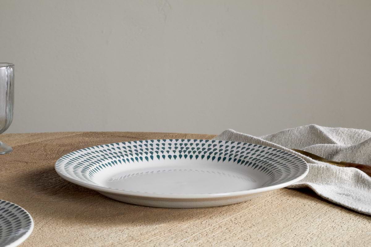 The nkuku Juniper Drop Dinnerware Set - Cream & Juniper (Set of 12) features a white ceramic plate with juniper green accents around the rim, displayed on a wooden table beside a beige napkin and clear glass.