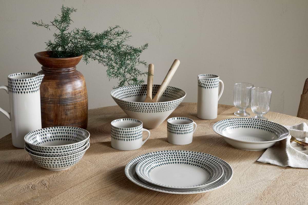 A set of nkuku Juniper Drop Dinner Plates in Cream & Juniper, featuring dark dotted patterns, is arranged on a wooden table with matching bowls, mugs, pitchers, glasses, and a brown vase with greenery in the background.