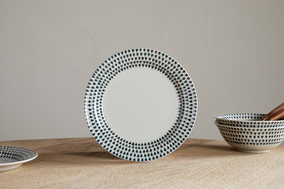 The nkuku Juniper Drop Dinnerware Set - Cream & Juniper (Set of 12) features a white ceramic plate with blue drop details on the rim, standing upright on wood, with matching bowls and a plate displayed nearby against a light wall.